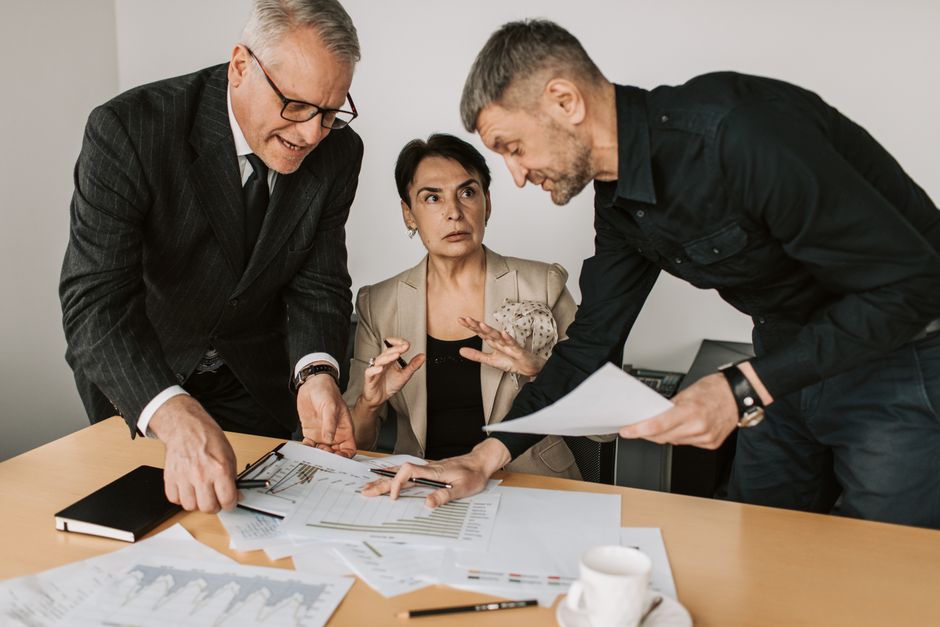 Three adults engaged in a dynamic discussion at an office table, analyzing documents and planning strategies.